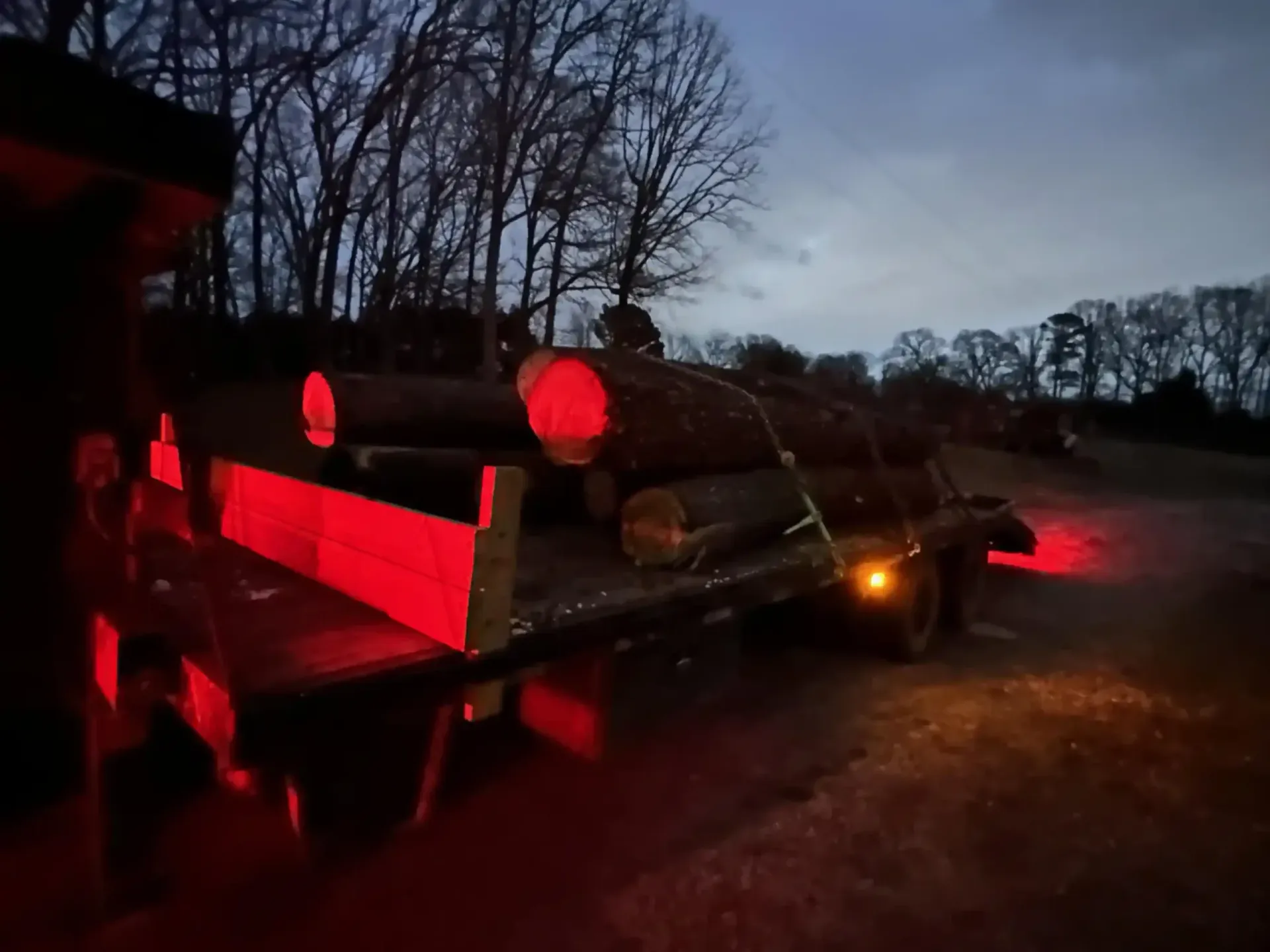 Red-lit trailer carrying logs on a dirt road at dusk, with headlights visible ahead.