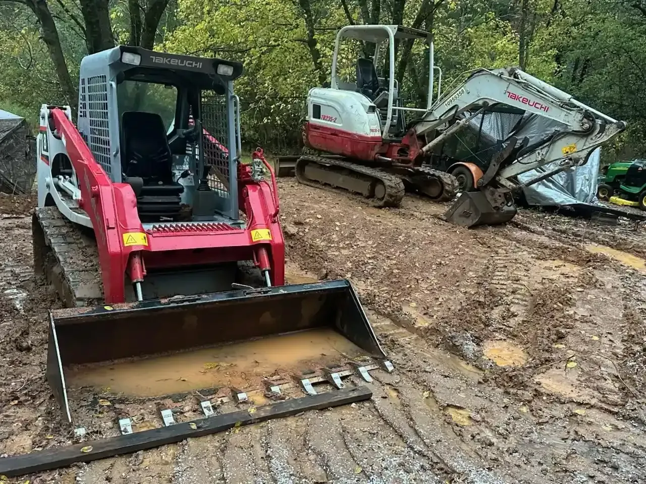 Two compact excavators on a muddy trail, one red and one white, with a bucket in the foreground.