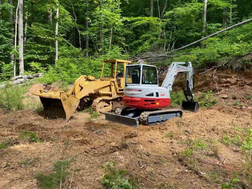 Yellow bulldozer and white excavator working on a dirt lot in a wooded area