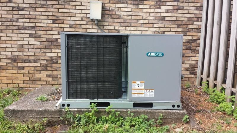 A gray outdoor HVAC unit sits on a concrete slab against a brick wall next to a wooden fence.