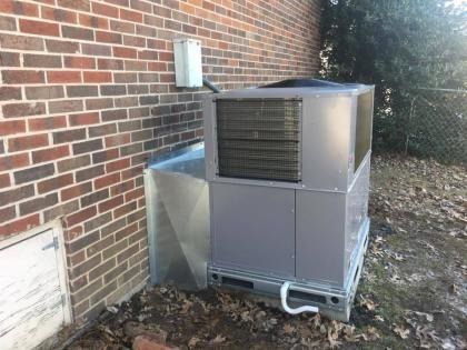 A grey HVAC packaged unit installed outside a brick building, connected to the wall with a galvanized metal duct.