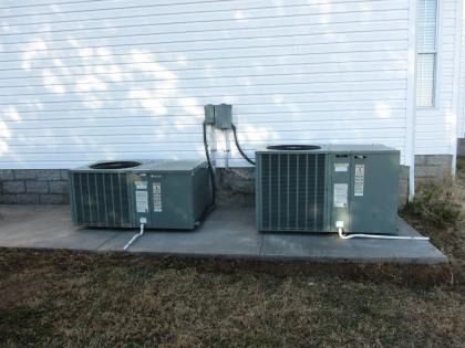 Two gray HVAC condenser units sit on a concrete slab next to a white-sided house.