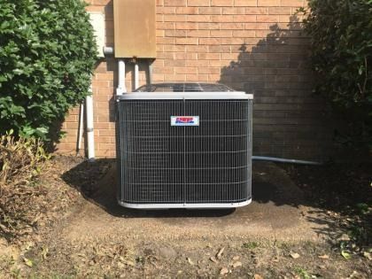 A grey central air conditioning unit sits on a concrete pad against a brick wall, surrounded by bushes.