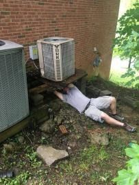 A person lies on the ground under a makeshift wooden stand supporting an outdoor air conditioning unit next to a wall.