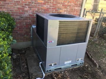 A gray HVAC unit sits on a concrete pad next to a red brick wall, with a small bush nearby.