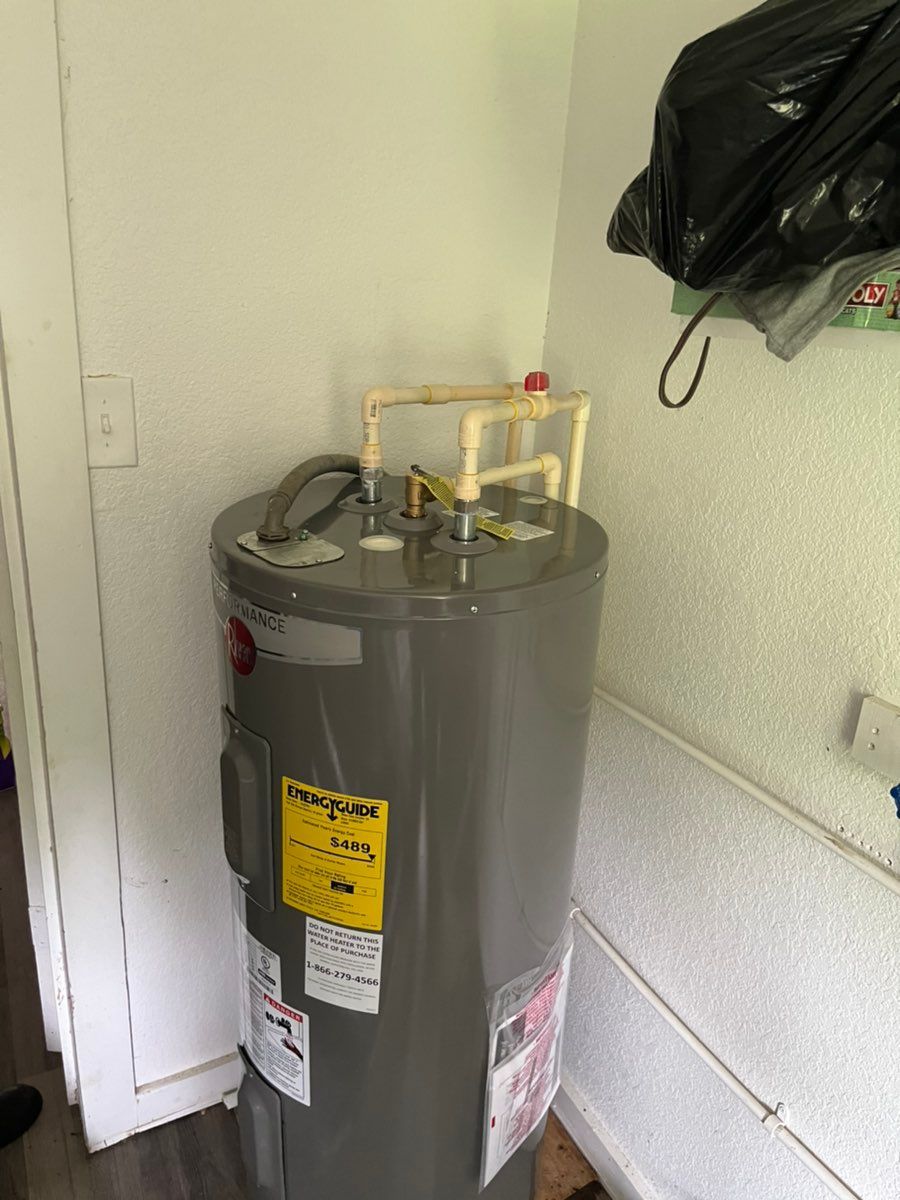 Gray water heater in a utility closet with pipes and a black bag on the wall.