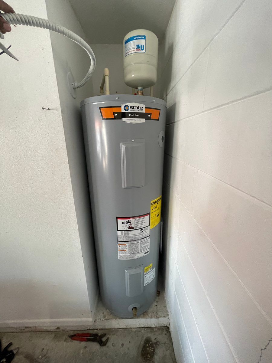 Gray water heater in a utility closet beside white walls and a flexible hose, with a small tank above it