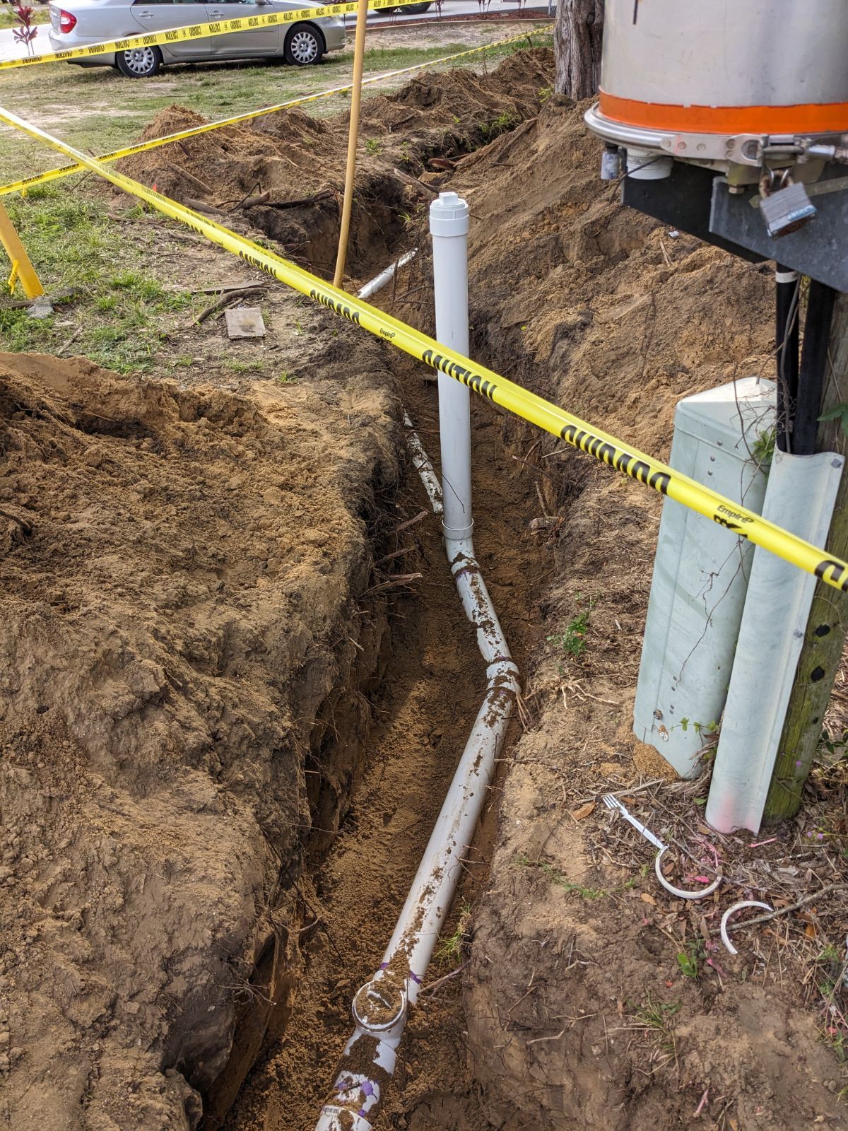 Trenching beside a driveway with exposed white pipes and yellow caution tape near a road.