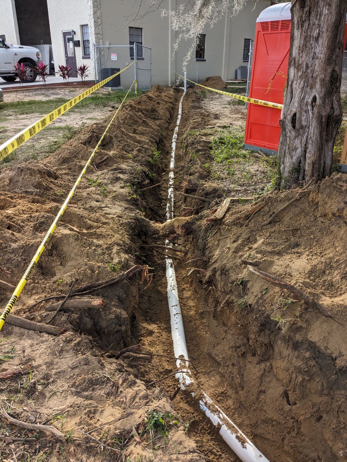 Trench dug along a sidewalk with white pipe and orange barrier tape beside a tree and red utility box