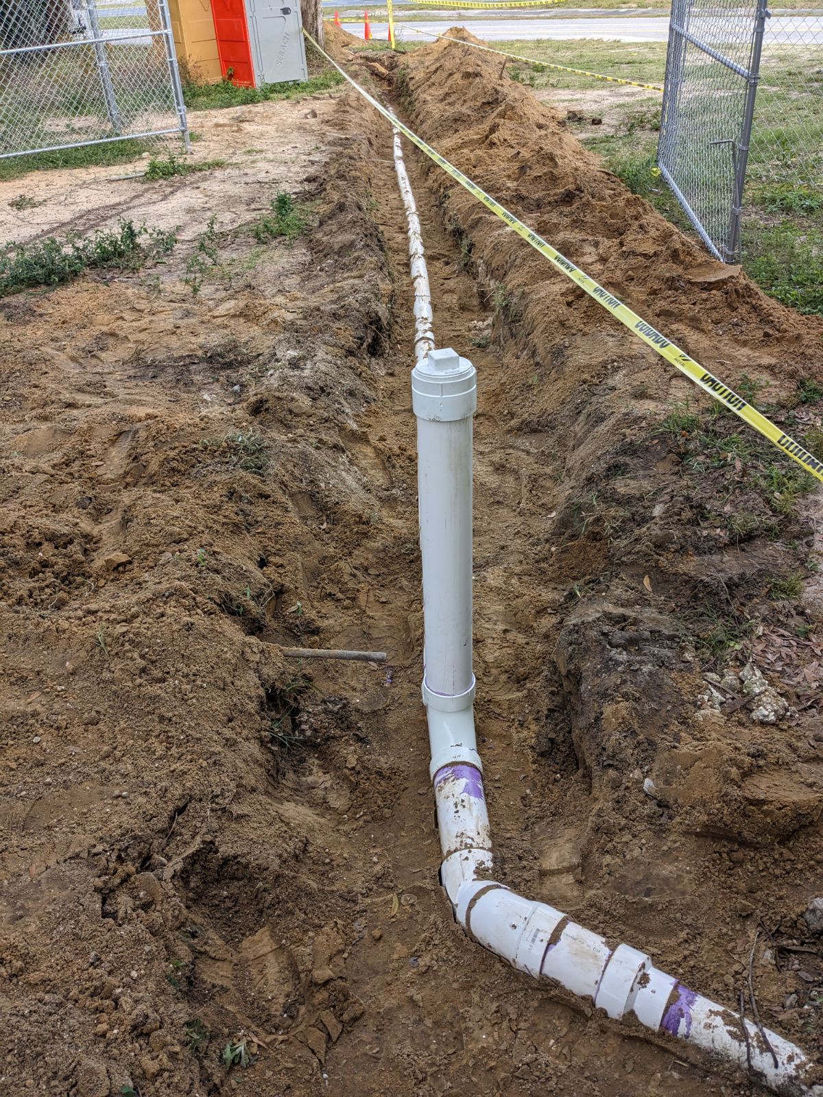 Underground white PVC pipe in a dug trench beside a fenced yard, with a vertical vent pipe and measuring tape.