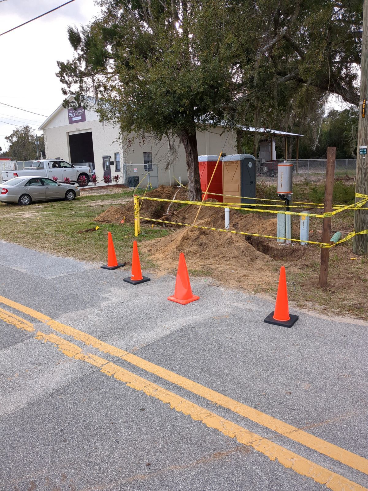 Orange cones and yellow caution tape block part of a roadside driveway near a tree and building.
