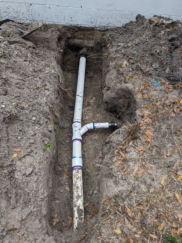 Excavated trench exposing a white PVC pipe and T-joint in muddy soil near a concrete wall