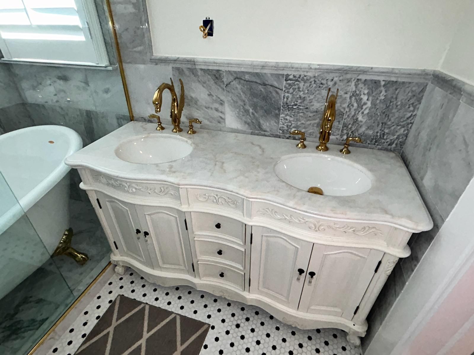 White marble double-sink vanity with gold faucets in a bathroom, beside a glass shower and tiled floor