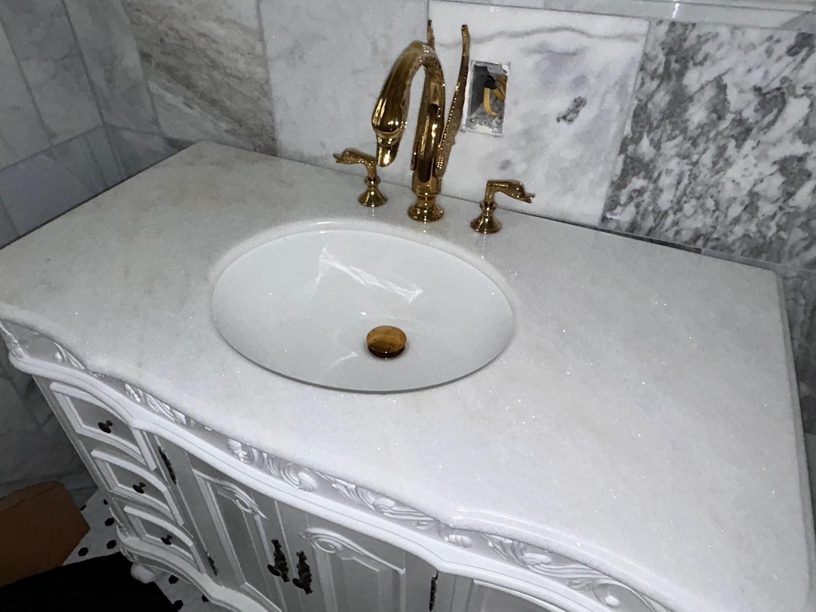 White bathroom vanity with oval sink and gold faucet against marble tile walls