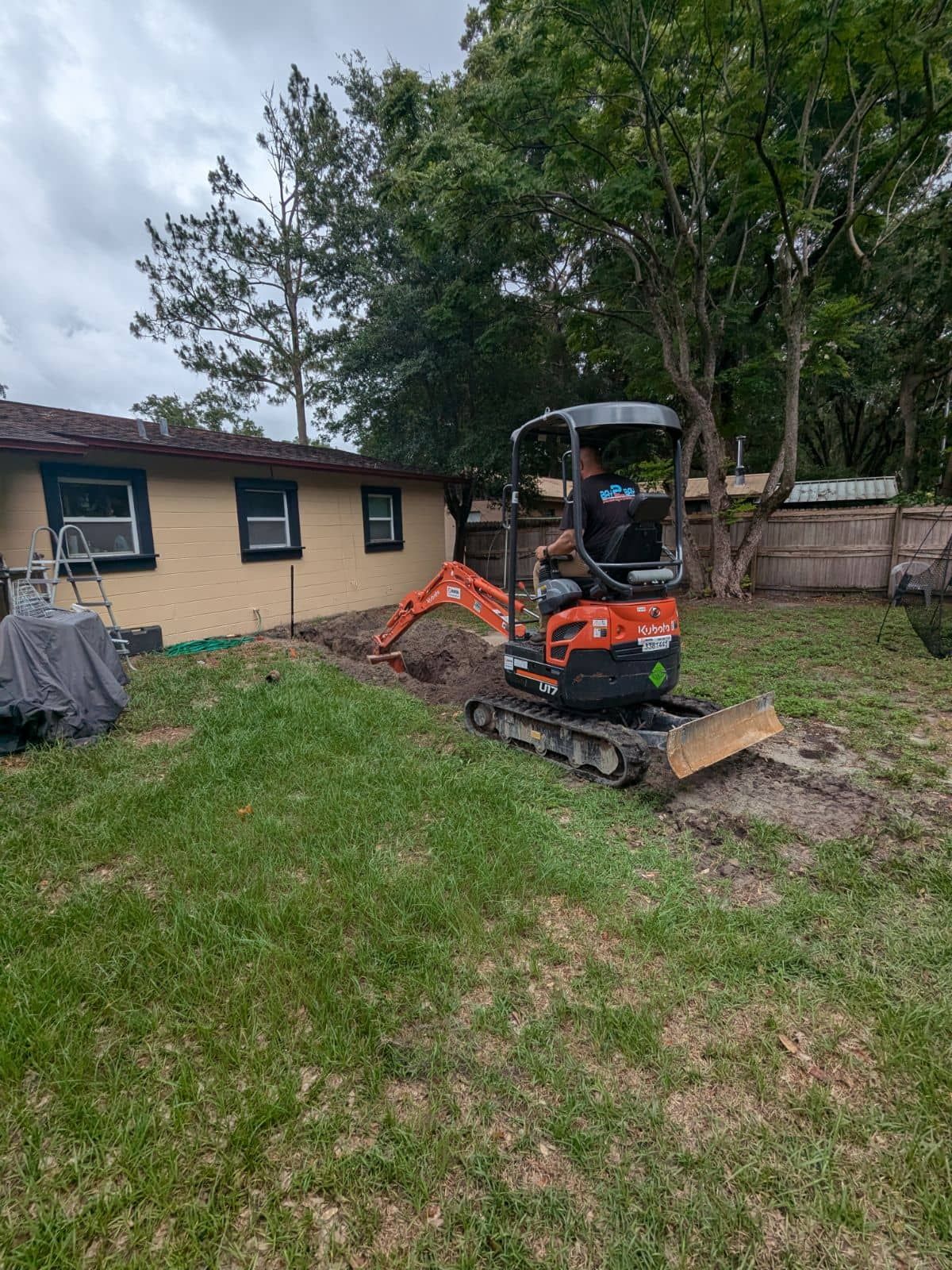Small orange excavator on a grassy yard beside a house and trees, digging near a muddy patch