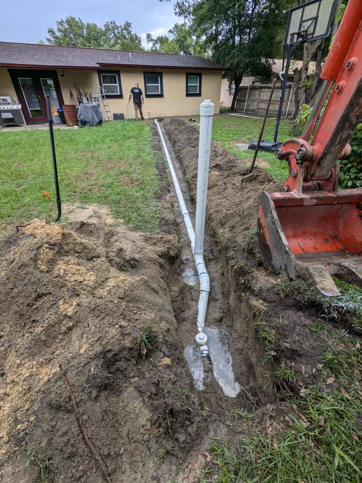 PVC drain pipe trench beside a house, with an excavator and dirt pile in the yard