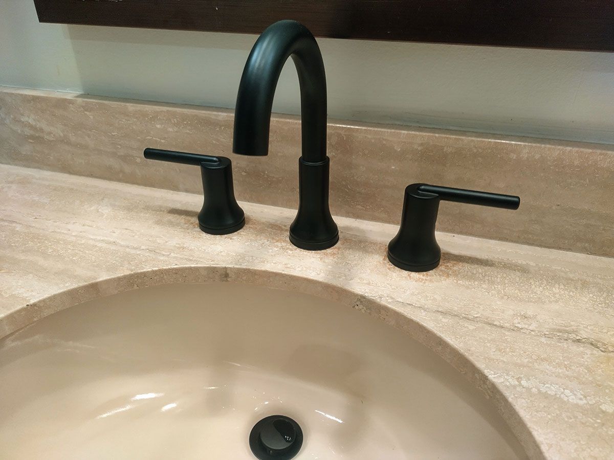 Bathroom sink with matte black faucet and handles against a beige countertop and backsplash.