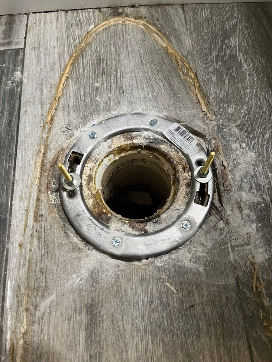 Circular metal floor flange around a dark opening, with two bolts and a yellow strap on a wood surface