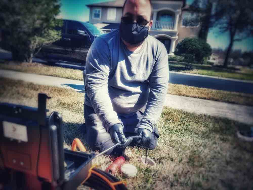 Person in gray shirt and mask kneels by a front yard fire pit, tending a small fire with tools.