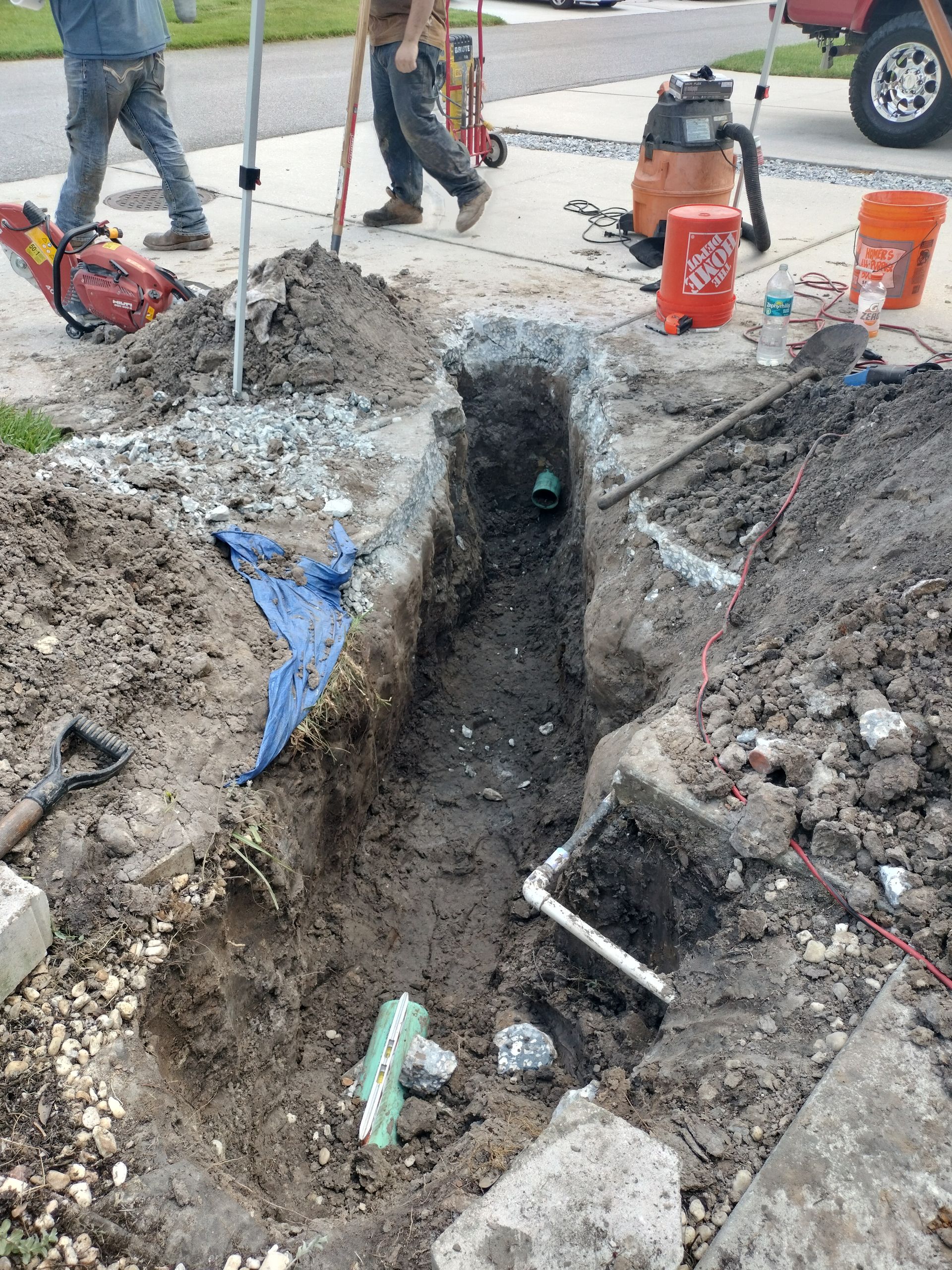Workers stand by a deep trench with dirt piles and orange buckets at a sidewalk construction site.