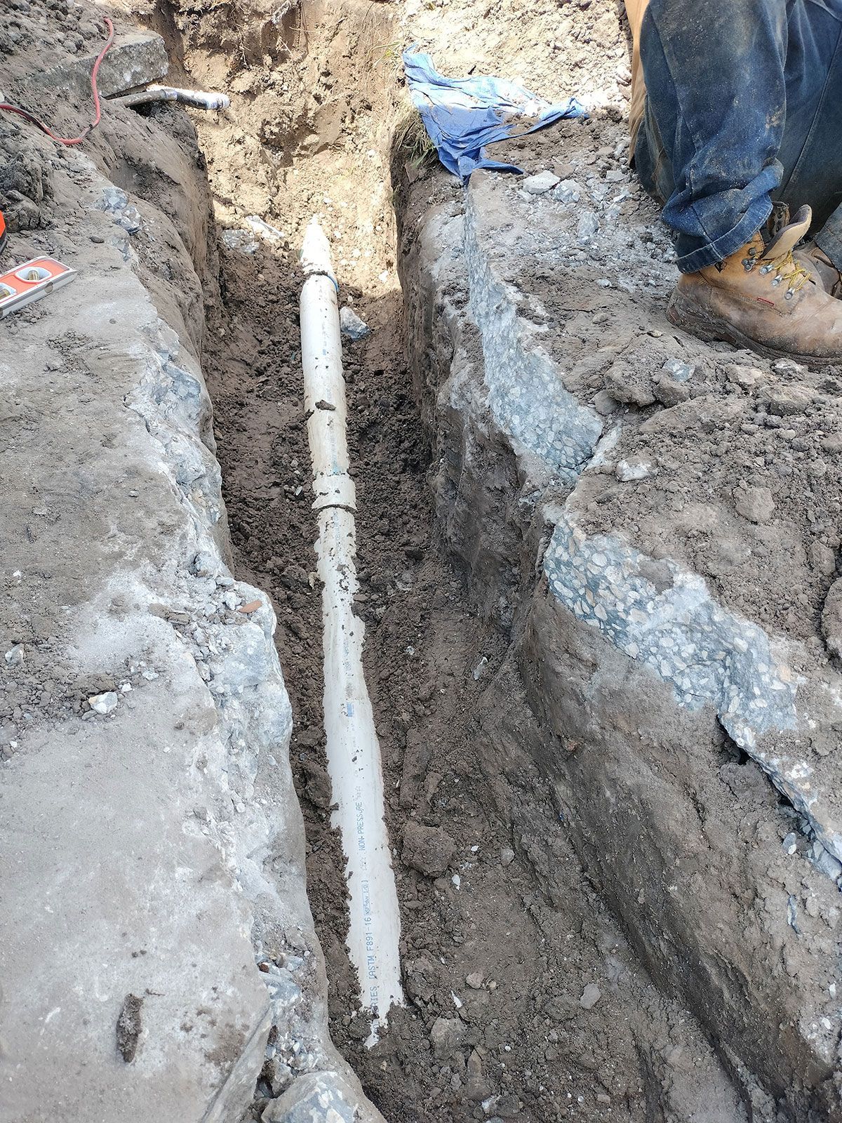 Trenched ground with a white PVC pipe laid in mud beside a worker’s boot