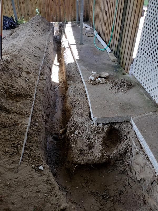 Backyard trench beside a concrete walkway, with a deep excavation along a fence line.