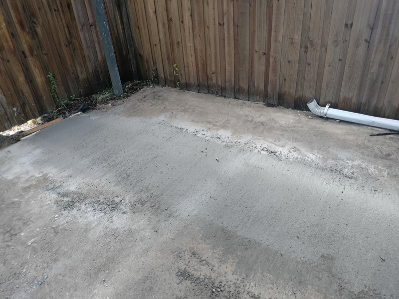 Concrete patio corner with a wooden fence and a white pipe along the wall, showing some stains and wear