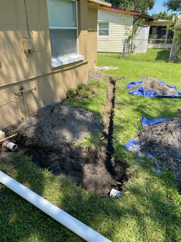 Backyard trench beside a beige house, with soil piles, white pipe, and blue tarp on grass