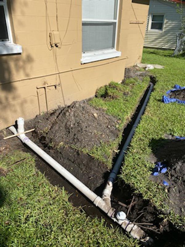 Black drainage pipe and white PVC pipes beside a tan house, next to a dug trench and dirt pile.