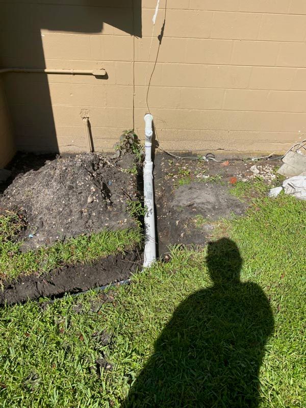Backyard by tan wall with a white pipe, mulch bed, grass, and a person's shadow in sunlight