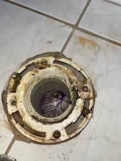 Old rusted toilet flange set in white tiled floor, with exposed pipe opening.