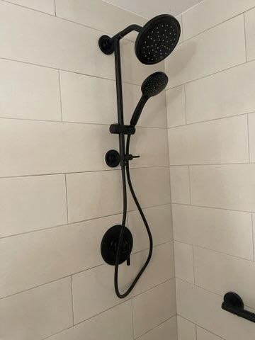 Black shower fixture with handheld sprayer and round overhead shower in tiled corner bathroom.