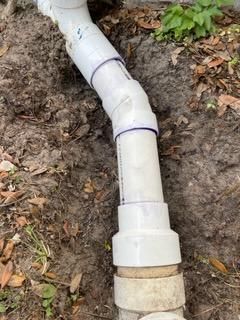 White PVC drainpipe with elbow joint emerging from soil and connecting to a larger pipe fitting outdoors
