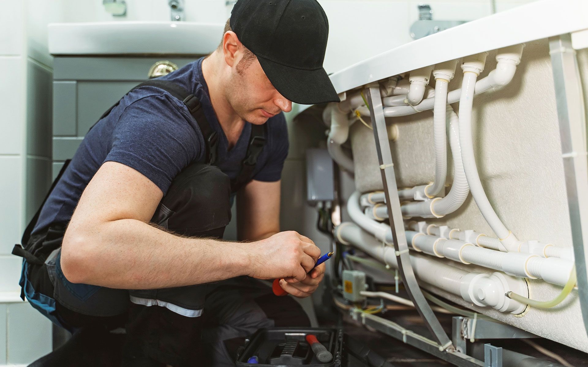 Technician crouches under a sink, repairing plumbing with tools in a kitchen or utility room.