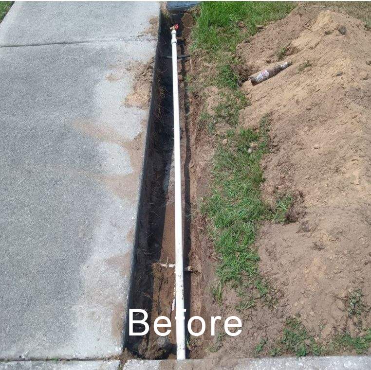 Narrow trench beside a concrete sidewalk, with a white pipe running along it and dirt piled on one side.