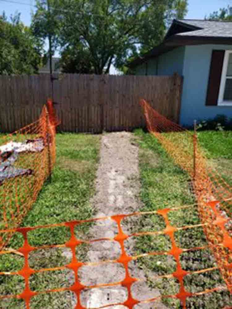 Narrow dirt path bordered by orange safety fencing leading through a backyard to a wooden fence and house.