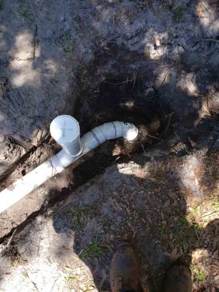 White PVC pipes and a capped pipe elbow on dirt in a shaded outdoor area.