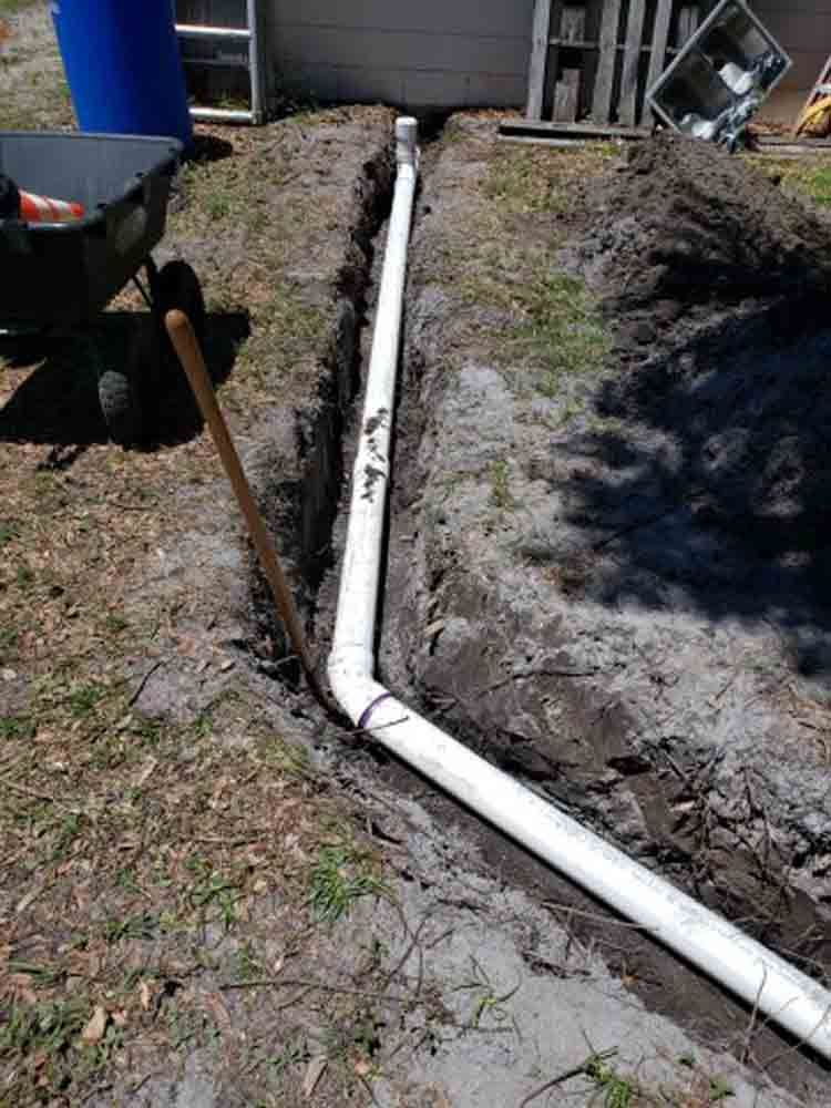 White PVC drainage pipe trench along a yard beside a house foundation and dirt slope.