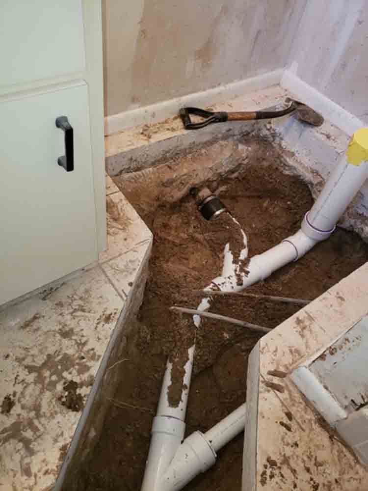 Bathroom floor cut open for plumbing repair, with exposed dirt and white PVC pipes.