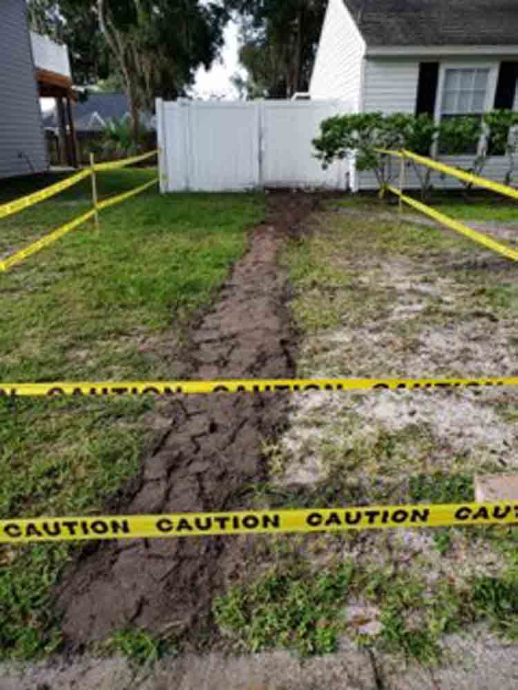 Yellow caution tape blocks a muddy path through a damaged yard toward a white fence and house