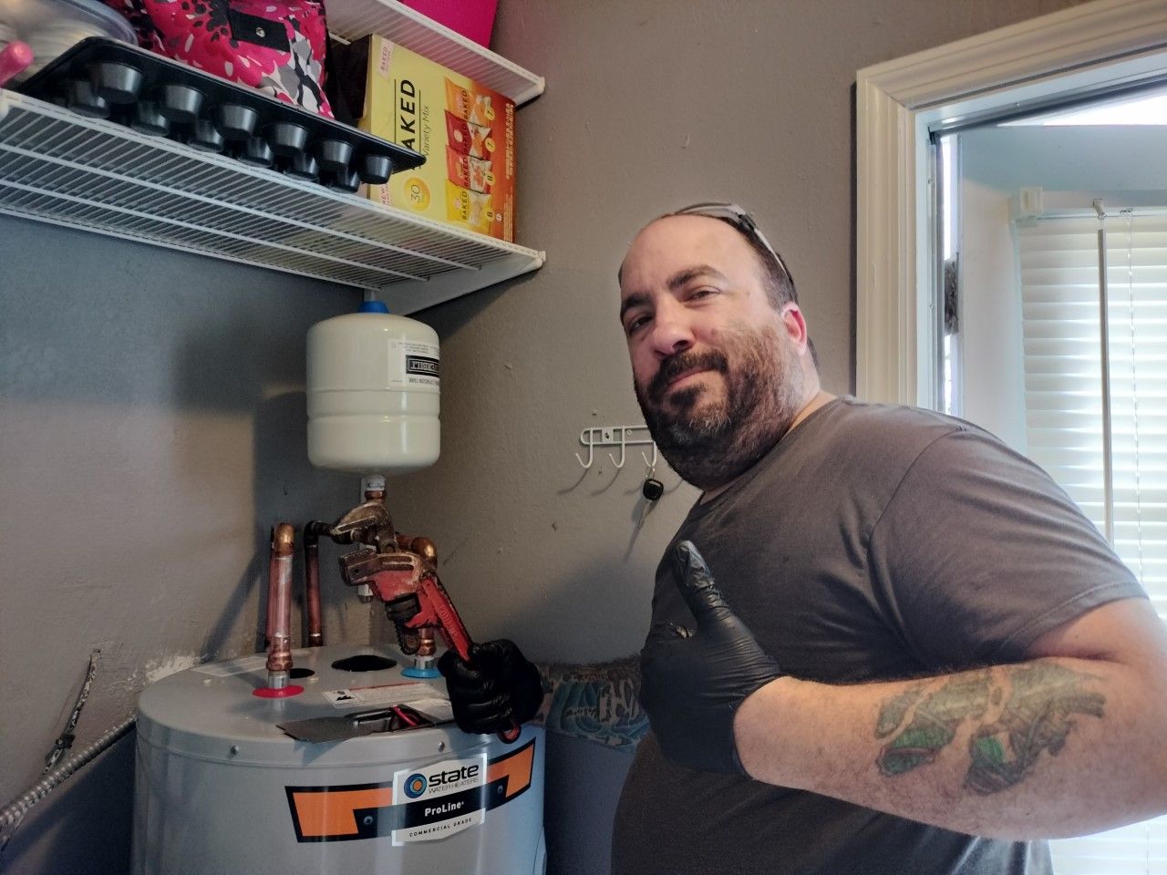 Man smiling beside a water heater, holding red plumbing tools in a small utility room