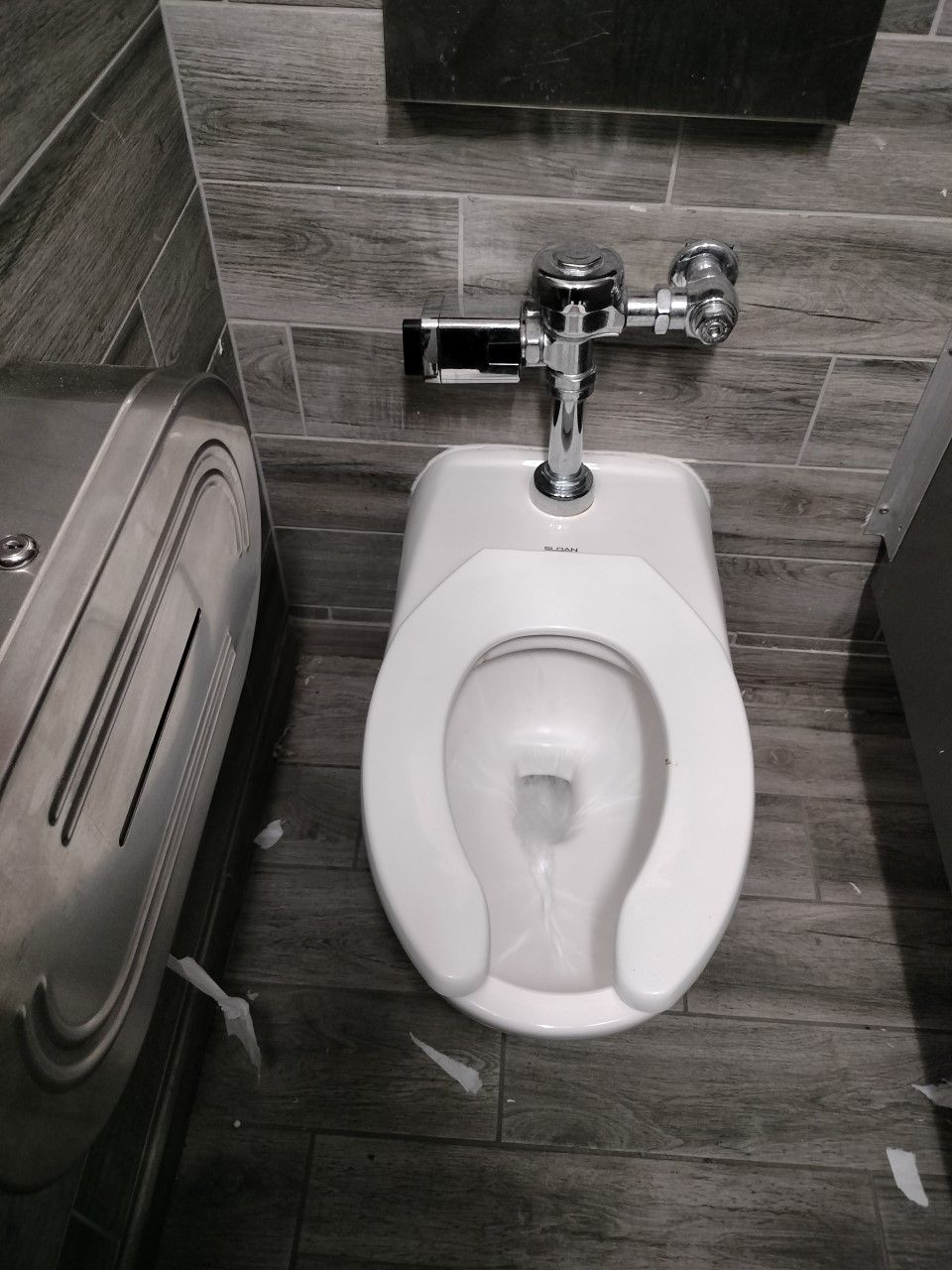Public restroom with a white toilet and chrome flush valve against gray tile walls.