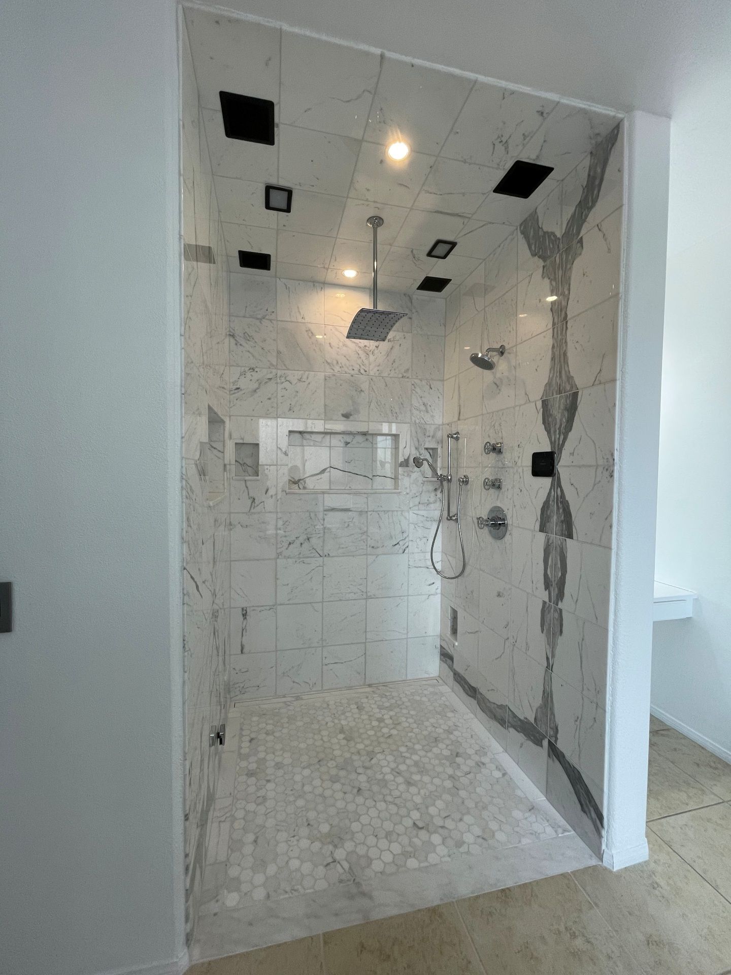 A walk-in shower with white marble-patterned tile walls, a pebble-stone floor, a rain showerhead, and recessed lighting.