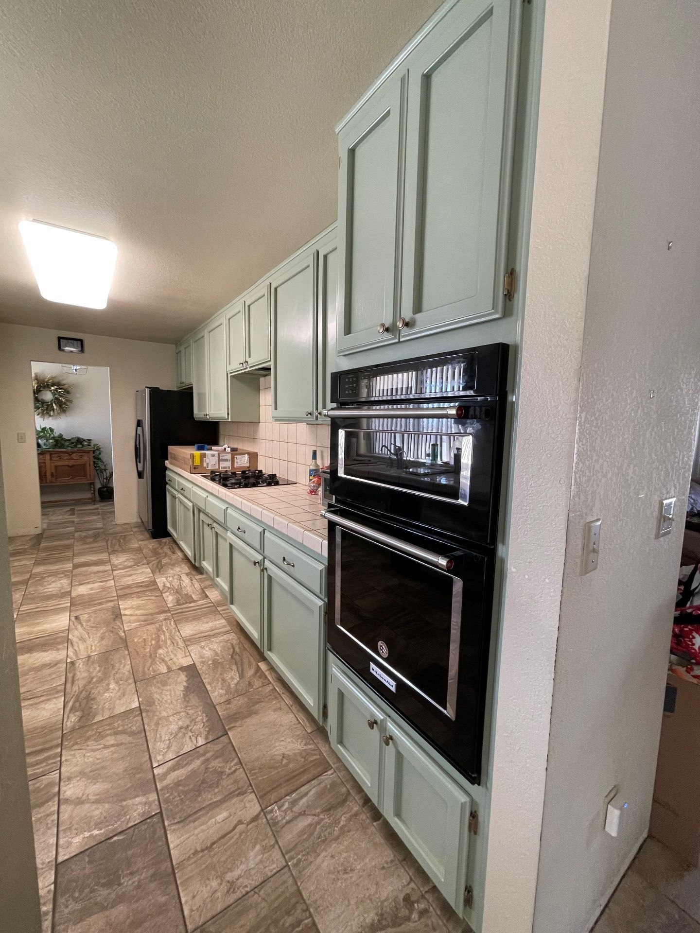 A kitchen with light green cabinets, a built-in black double oven, and brown patterned tile floors.
