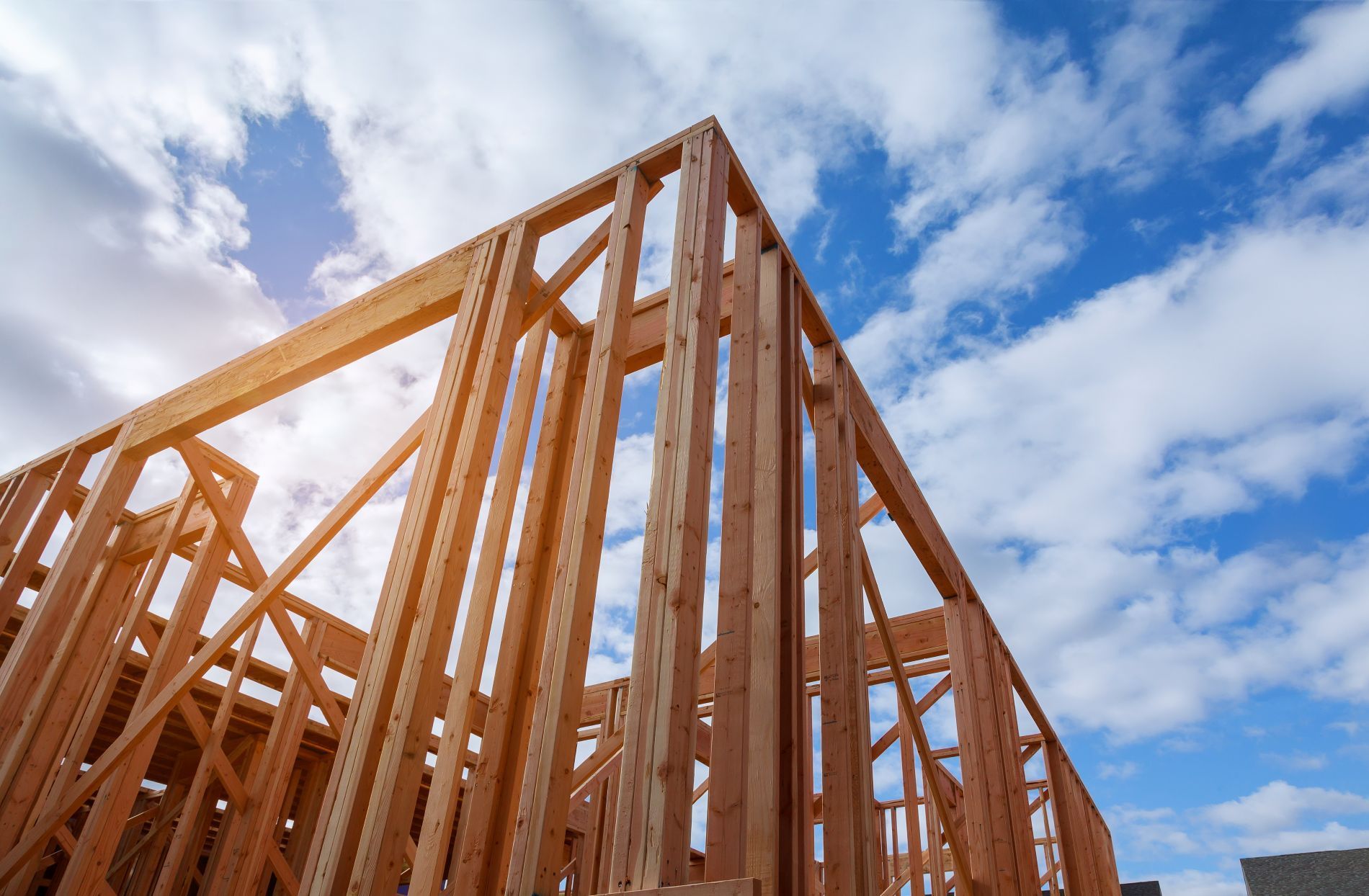 A low-angle view of a wooden house frame under a bright blue sky with white clouds.