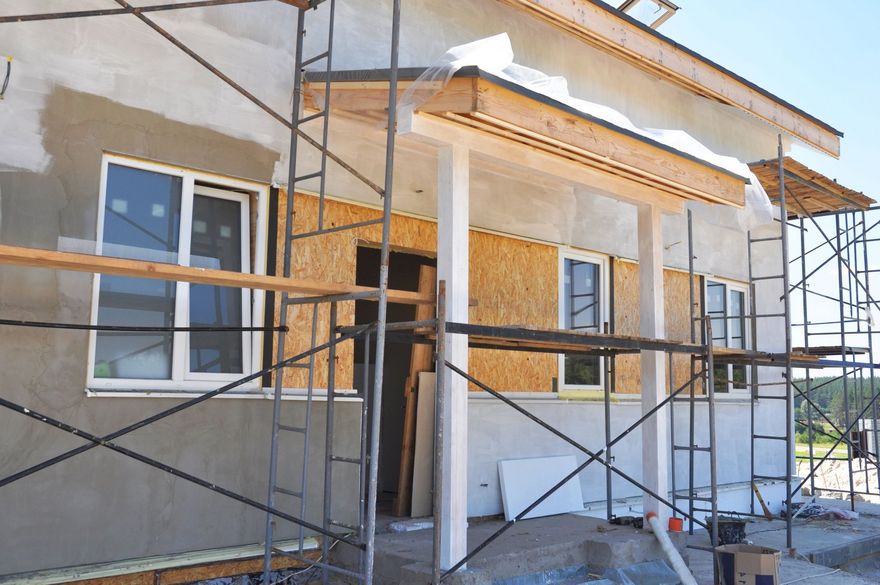 Exterior of a house under construction featuring partial plaster walls, exposed wooden siding, and metal scaffolding.