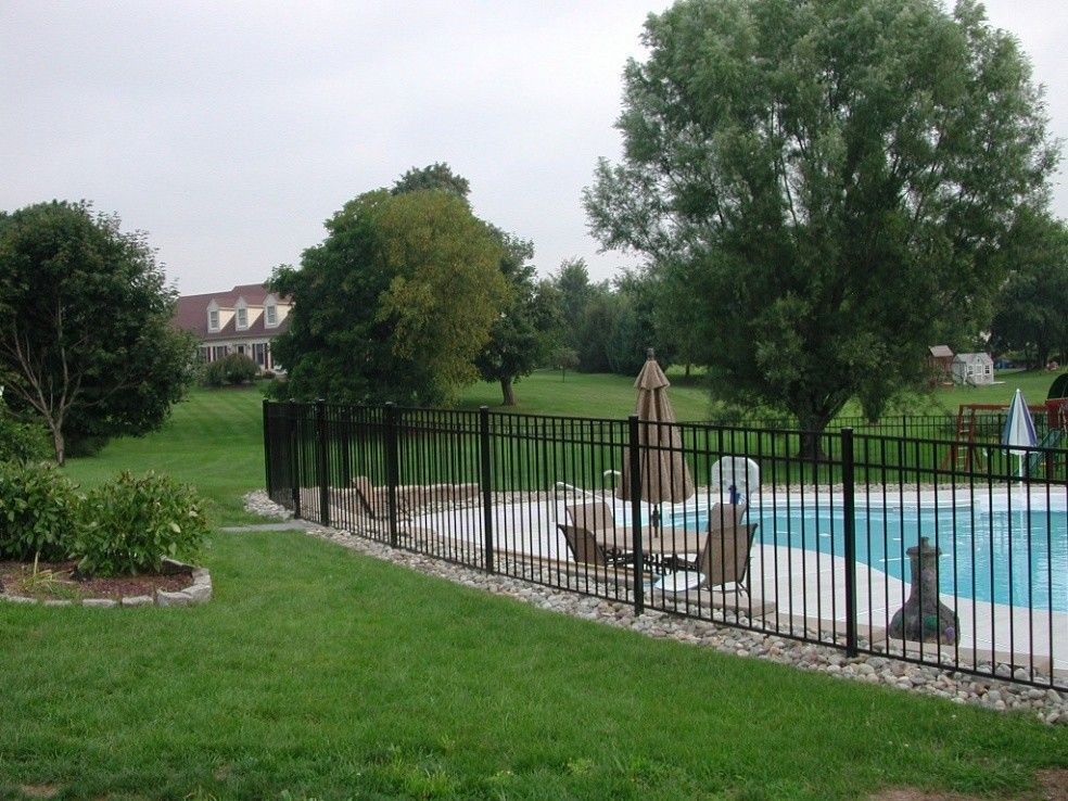 Black fence surrounds a swimming pool in a green yard with trees and a house in the background. Black fence surrounds a swimming pool in a green yard with trees and a house in the background.