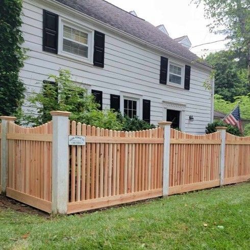 Cedar picket fence in front of a white house with black shutters; American flag in the background.