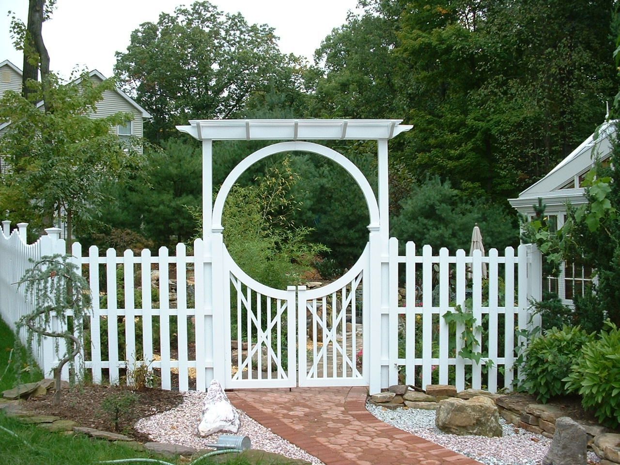 White picket fence with a round-arched gate, leading to a brick path in a garden.
