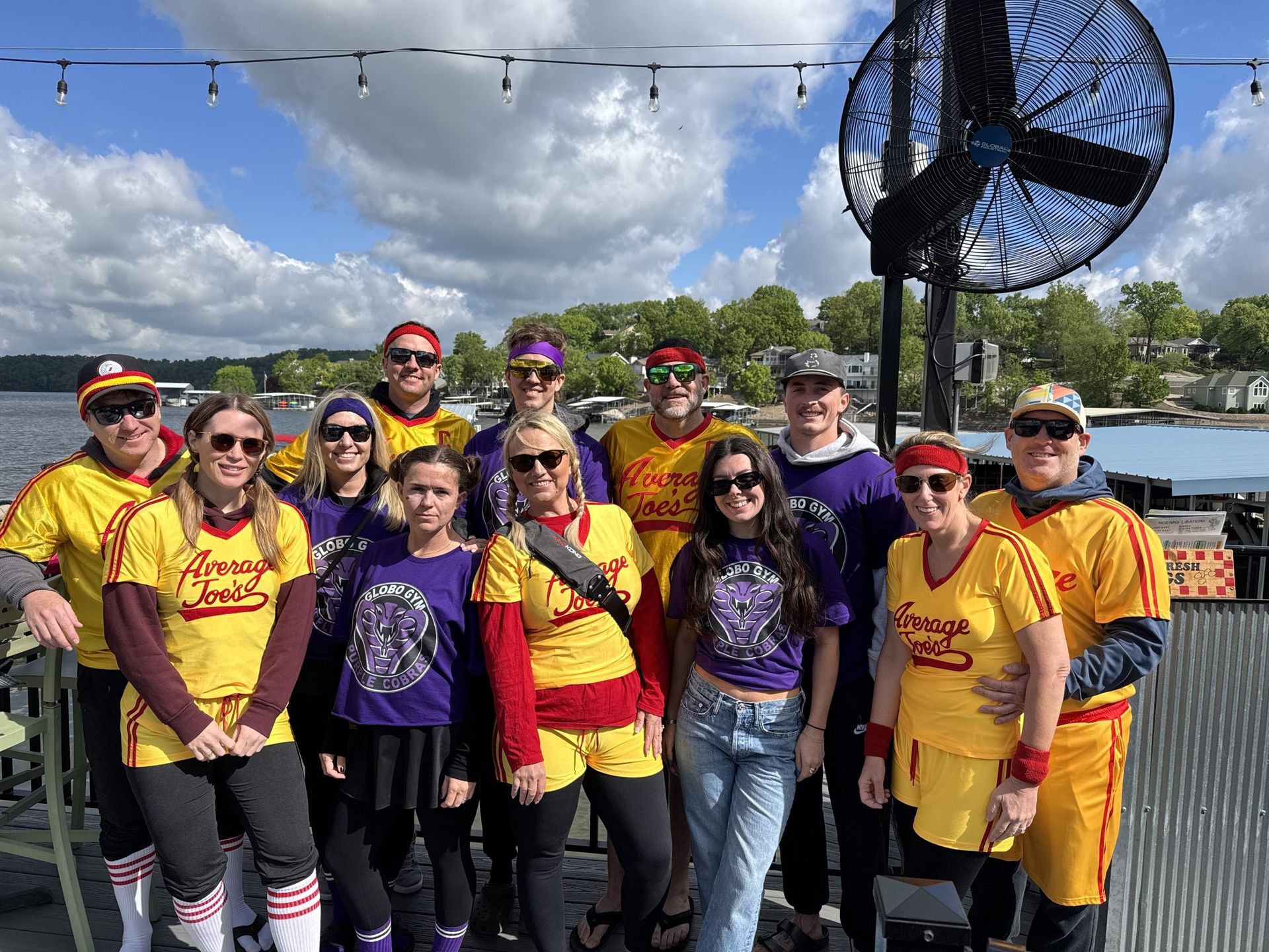 Group in purple and yellow shirts posing outdoors by the water under a cloudy sky
